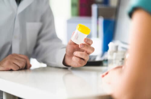 Image of two people sitting at a desk, one is handing the other a cup for a urine test.